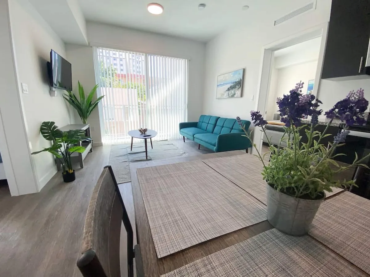 The image showcases a bright, open-plan living and dining area featuring a stylish teal sofa, a wall-mounted TV for relaxation, and a contemporary wooden dining table set with chic placemats and a potted lavender centerpiece. A large sliding glass door fills the space with natural Miami sunlight and offers easy access to outdoor views, creating a calm and inviting atmosphere ideal for unwinding or socializing.