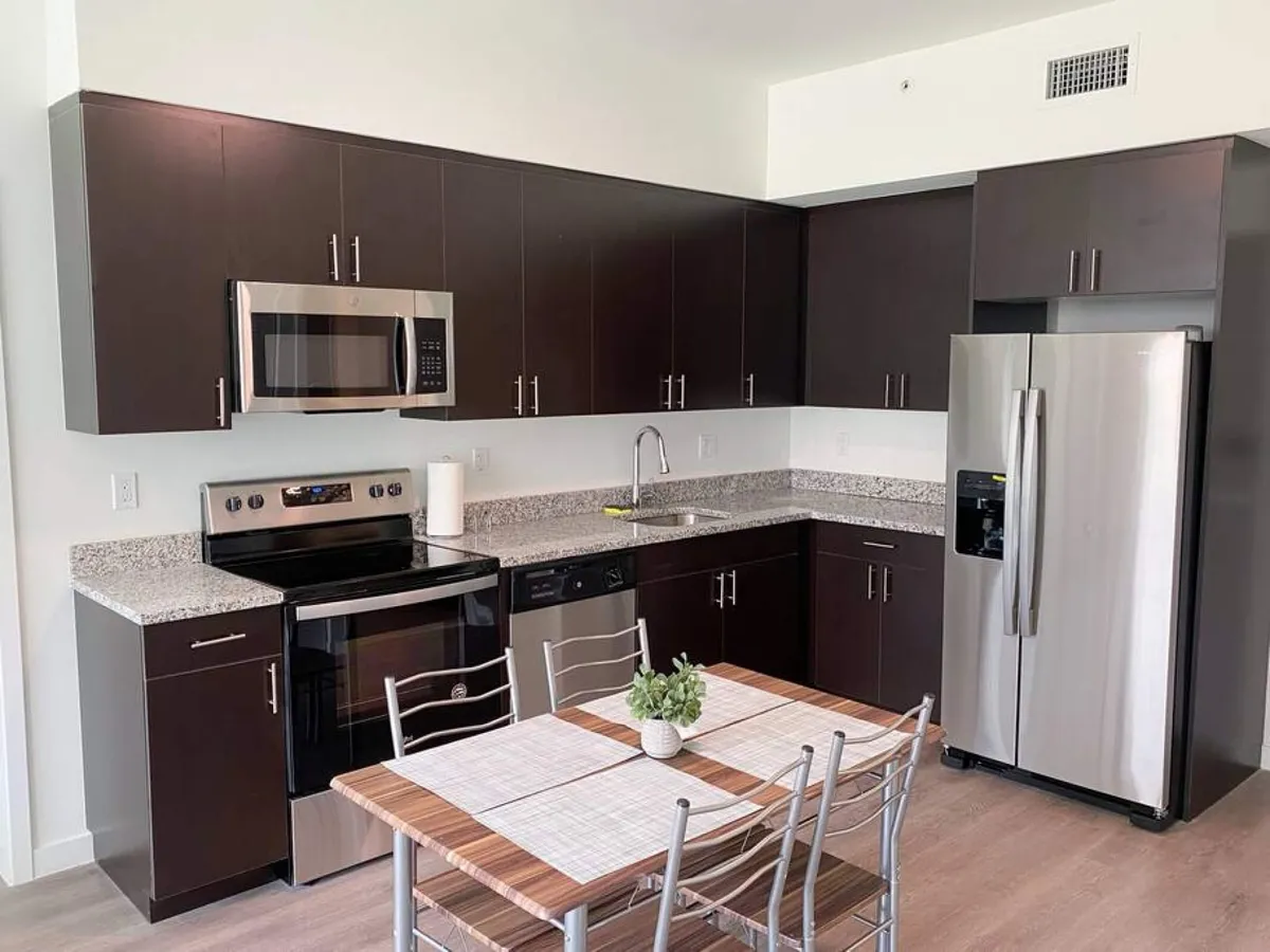 This image presents a spacious kitchen featuring stainless steel appliances, including a refrigerator, oven, and microwave. The L-shaped dark wood cabinetry adds warmth and sophistication, while the granite countertops provide ample workspace for meal prep. In the foreground, a large wooden dining table with metal chairs and simple placemats is ready for gatherings, offering a seamless blend of cooking and dining areas.