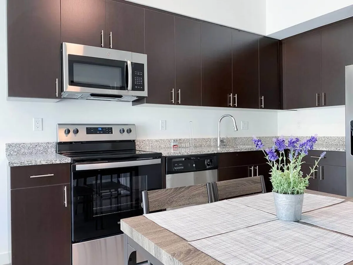 A different angle of the kitchen focuses on the stainless steel oven and microwave, complemented by dark wood cabinetry and a granite countertop. The dining table is set with a lavender floral centerpiece, adding a touch of elegance and homeliness to the space. The overall ambiance is modern, fresh, and inviting, reinforcing the kitchen's role as a hub for connection and creativity.