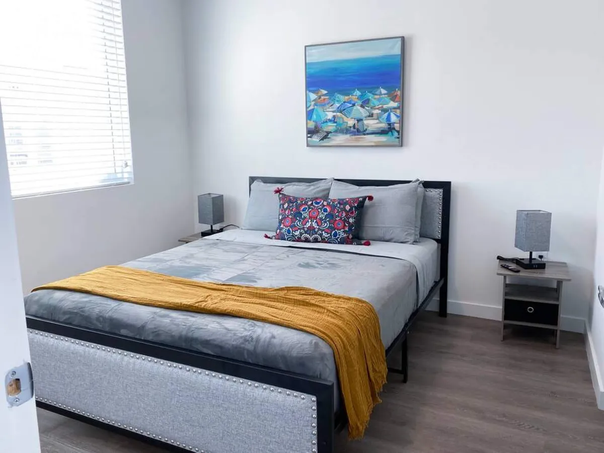 A serene bedroom with a queen-sized bed dressed in grey bedding and a mustard throw, modern bedside tables and lamps, and a vibrant beach painting above the bed. The space is filled with natural light, offering a calm and restful retreat.