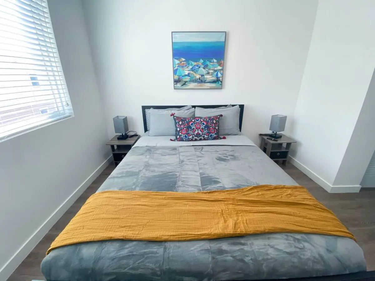 This image showcases a serene bedroom featuring a plush queen bed adorned with grey velvet bedding and a mustard yellow throw. The room is accented by a decorative pillow, matching bedside tables with lamps, and calming beach-themed wall art. The overall ambiance is bright, cozy, and inviting, emphasizing comfort and style.