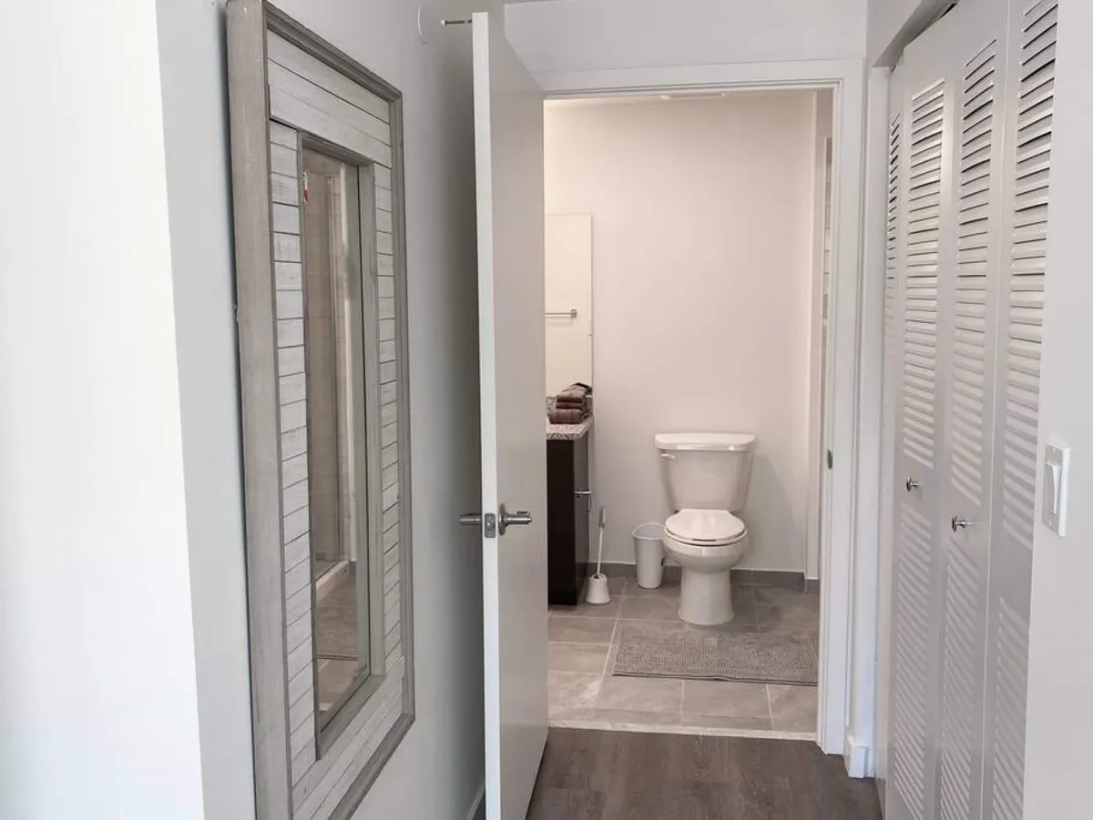 The image features an open door revealing a clean bathroom, flanked by a large wall mirror with a rustic frame and white louvered closet doors. The area is tidy, fresh, and functional, evoking a sense of order and readiness upon arrival.