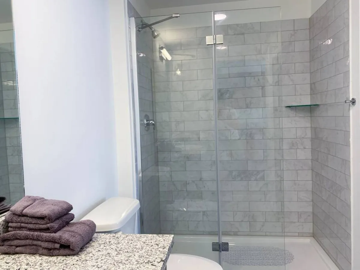 A modern ensuite bathroom featuring a sleek glass shower enclosure with tiled walls, a granite countertop with a sink, and a stack of folded purple towels. The space is clean, bright, and thoughtfully designed for relaxation and rejuvenation after a long day.