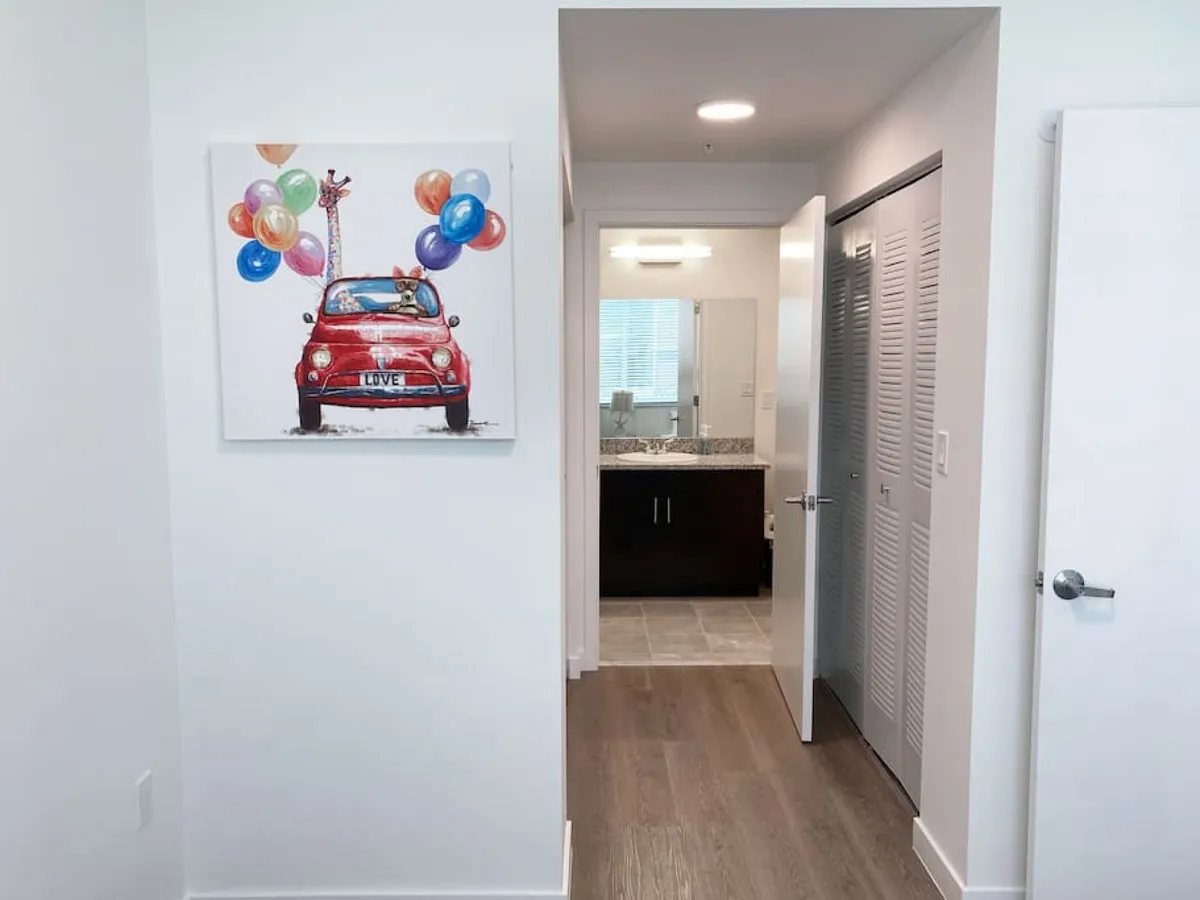 This image captures a welcoming interior hallway with a door leading to the bathroom, accented by modern lighting, wooden floors, and cheerful, colorful artwork featuring a red car with balloons and a giraffe. The scene is well-lit and gives a sense of transition from the entryway into the apartment’s living spaces.