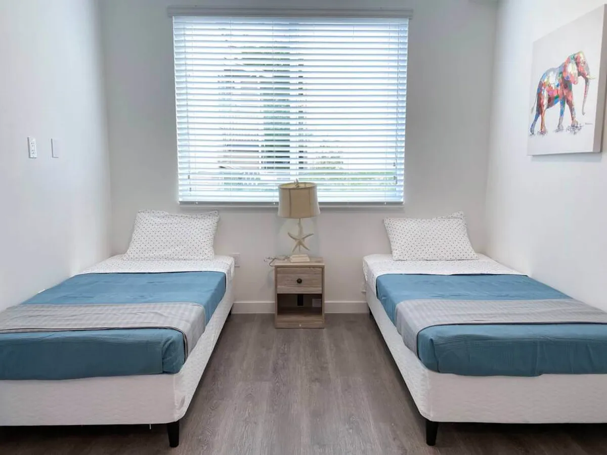 This image depicts a second bedroom with two single beds, each dressed in blue bedding. A nightstand with a starfish-base lamp sits between the beds, while a large window with white blinds lets in natural light. A colorful elephant painting adds a playful touch, creating a bright and airy atmosphere ideal for guests needing separate beds.
