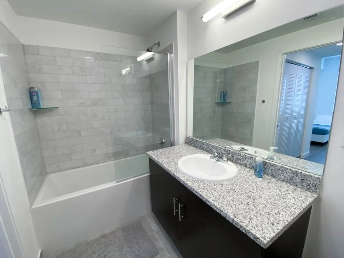This interior view centers on a granite countertop with sink, a bathtub with a glass shower screen, and a large mirror reflecting the entrance to the bedroom. The space is bright, practical, and inviting, emphasizing both function and relaxation.